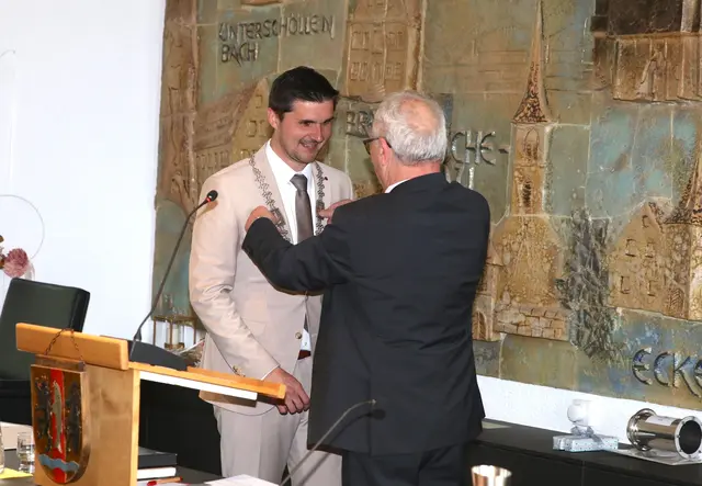 Zwei Männer. Links der Eckentaler Bürgermeister Sebastian Singer. Rechts daneben von hinten zu sehen Konrad Gubo. Er legt Sebastian Singer gerade die Amtskette des Bürgermeisters um.